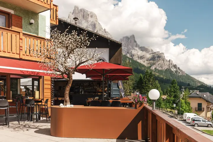 La Terrazza Montanara di giorno: l'albero nella sua fioriera, gli ombrelloni rossi, dietro le Pale di San Martino