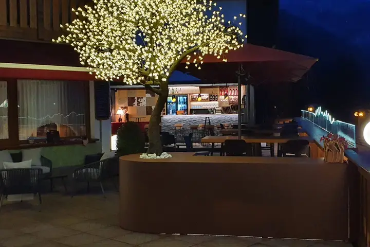 La stessa terrazza di sera, con l'albero illuminato e, dietro, il bar acceso